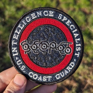 May include: A black and white embroidered patch with a red border. The patch says "U.S. Coast Guard Intelligence Specialist" around the outside and has a stylized octopus in the center.