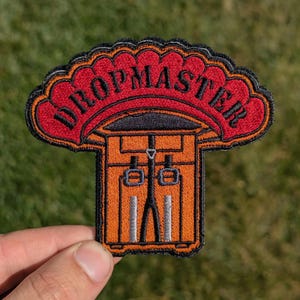 May include: An embroidered patch with a red parachute and the text "DROPMASTER" in black. The parachute is over a brown and black illustration of a parachute door with a harness.
