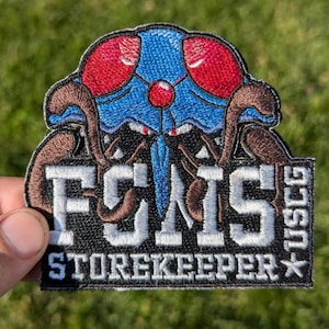 May include: Embroidered patch featuring a blue and red bug with large eyes and the text "FCMS STOREKEEPER -115CC".