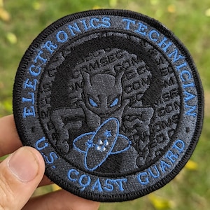 May include: A gray and blue embroidered patch with the text "U.S. Coast Guard Elite Electronics Technician" around the edge. The center of the patch features a stylized black creature with glowing eyes and a symbol of an atom.