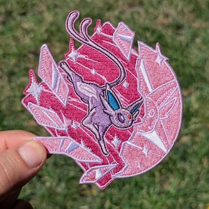 May include: An embroidered patch featuring a purple creature with long ears and a tail, positioned against a circular background of dark pink and light pink crystalline shards, accented with white star-shaped sparkles throughout the design.