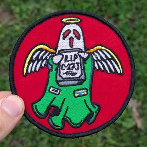 May include: A circular embroidered patch with a cartoon ghost design. The ghost has angel wings, a halo, and is holding a tombstone with the text "RIP C23". The patch is set on a red background with a black border and green details.