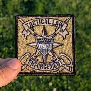 May include: Embroidered patch with the words "TACTICAL LAW ENFORCEMENT" and a starburst design. The patch features a shield with "USCG" in the center. The patch is square with a black border and a tan background.