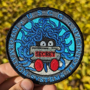 May include: A blue and gray embroidered patch with a cartoon monster holding a box that says "SECRET". The text around the monster reads "USCG Information Systems Technician".