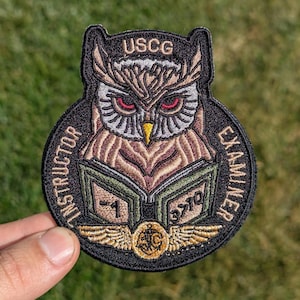 May include: A black and gold embroidered patch with an owl wearing a graduation cap and holding a book. The patch says "USCG Instructor Examiner" and has a gold eagle emblem.