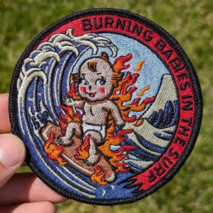 May include: A red, white, and blue embroidered patch with a cartoon baby riding a surfboard on a wave of fire. The text "Burning Babies in the Surf" is written around the edge of the patch.