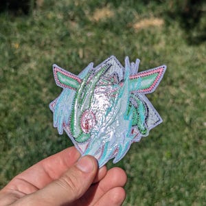 Op de afbeelding: Een decoratieve geborduurde patch met een gestileerd wezenontwerp met iriserende, lichtreflecterende draden in tinten mintgroen, lichtblauw en zachtroze, afgewerkt met een witte rand en ingewikkeld stikwerk over de gehele vorm.