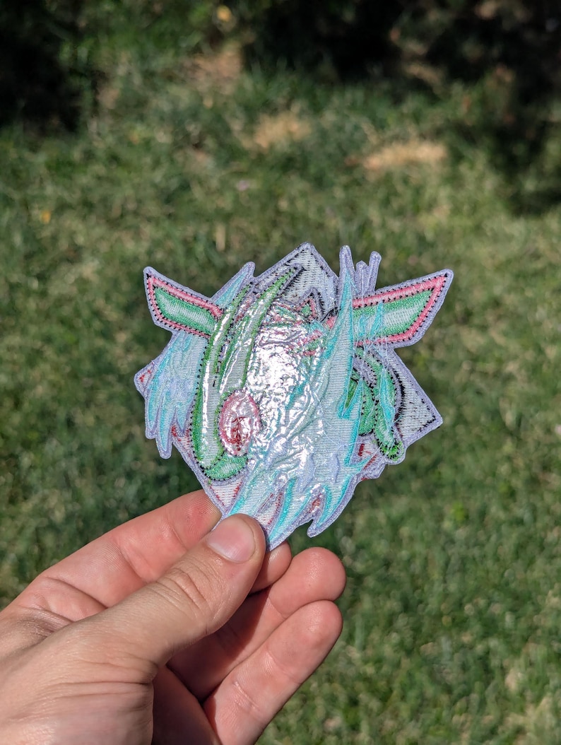 Op de afbeelding: Een decoratieve geborduurde patch met een gestileerd wezenontwerp met iriserende, lichtreflecterende draden in tinten mintgroen, lichtblauw en zachtroze, afgewerkt met een witte rand en ingewikkeld stikwerk over de gehele vorm.