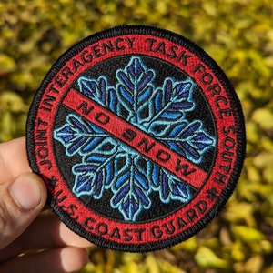 May include: A black and red embroidered patch with a blue snowflake crossed out with a red line and the text "NO SNOW". The text around the snowflake reads "Joint Interagency Task Force South US Coast Guard".