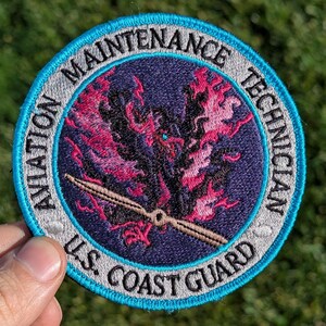 May include: A blue and gray embroidered patch with the text "Aviation Maintenance Technician U.S. Coast Guard". The patch features a stylized image of a propeller and flames.