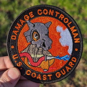 May include: Round patch with the text "DAMAGE CONTROLMAN U.S. COAST GUARD" in orange lettering on a black border. The patch features a cartoon character in a firefighter's uniform, with a gray skull-like head and a hose spraying blue water.