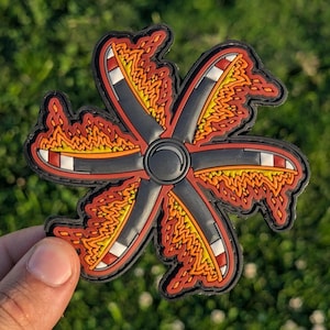 May include: A black, red, and orange patch with a unique design. The patch features a central black circle with six radiating arms. Each arm has a black base, red and white stripes, and orange flame-like details. The patch is held against a green background.