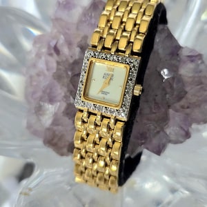 Puede incluir: Un reloj de pulsera Anne Klein dorado con esfera cuadrada y detalles de diamantes. El reloj presenta una correa dorada y una esfera crema con agujas y números romanos dorados. El reloj se muestra sobre una superficie negra.