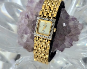 May include: A gold-toned Anne Klein wristwatch with a square face and diamond accents. The watch features a gold band and a cream-colored watch face with gold hands and Roman numerals. The watch is displayed on a black surface.