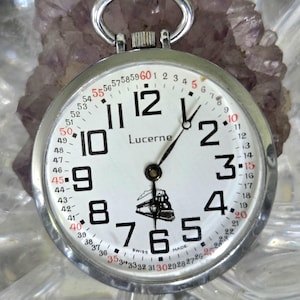 May include: A silver Lucerne pocket watch with a white face, black numerals, and a train graphic. The watch has a second hand and a ring for attaching a chain. The watch is marked "Swiss Made".