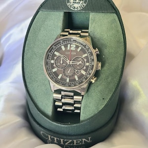 May include: A silver Citizen Eco-Drive chronograph watch with a black face and red accents. The watch is in a green box with the Citizen logo.