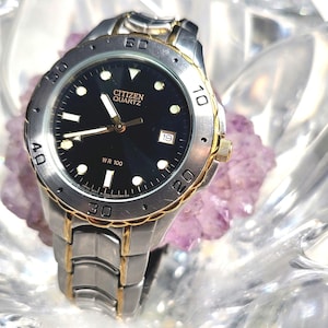 May include: A silver and gold Citizen Quartz wristwatch with a black face and gold accents. The watch has a date window and is water resistant to 100 meters.