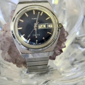 Könnte beinhalten: Eine silberne Timex Electric Uhr mit schwarzem Zifferblatt und silbernem Metallband. Das Zifferblatt der Uhr hat goldfarbene Stunden- und Minutenzeiger, einen goldfarbenen Sekundenzeiger und ein Datumsfenster, das "TUE 17" anzeigt.