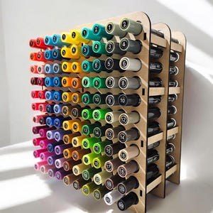 May include: A wooden storage rack for markers, with 98 slots, numbered 1 to 98. The rack is made of light brown wood and has three tiers. The markers are arranged in rows, with the caps facing out. The markers are a variety of colours, including red, orange, yellow, green, blue, purple, pink, brown, and black.