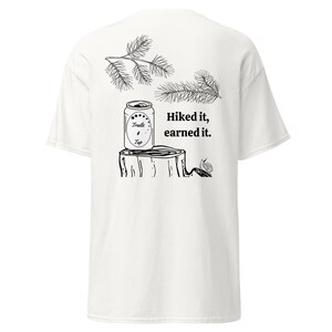 May include: White t-shirt with a black and white illustration of a can of "Trails & Tap" beer on a tree stump with a snail. The text "Hiked it, earned it." is printed on the shirt.