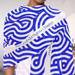 May include: A blue and white patterned scarf with the text "Nu Lambda Sigma Chapter" in white. The scarf features a repeating wave design in blue and white, creating a visually striking pattern. The scarf is draped over a person's shoulders.