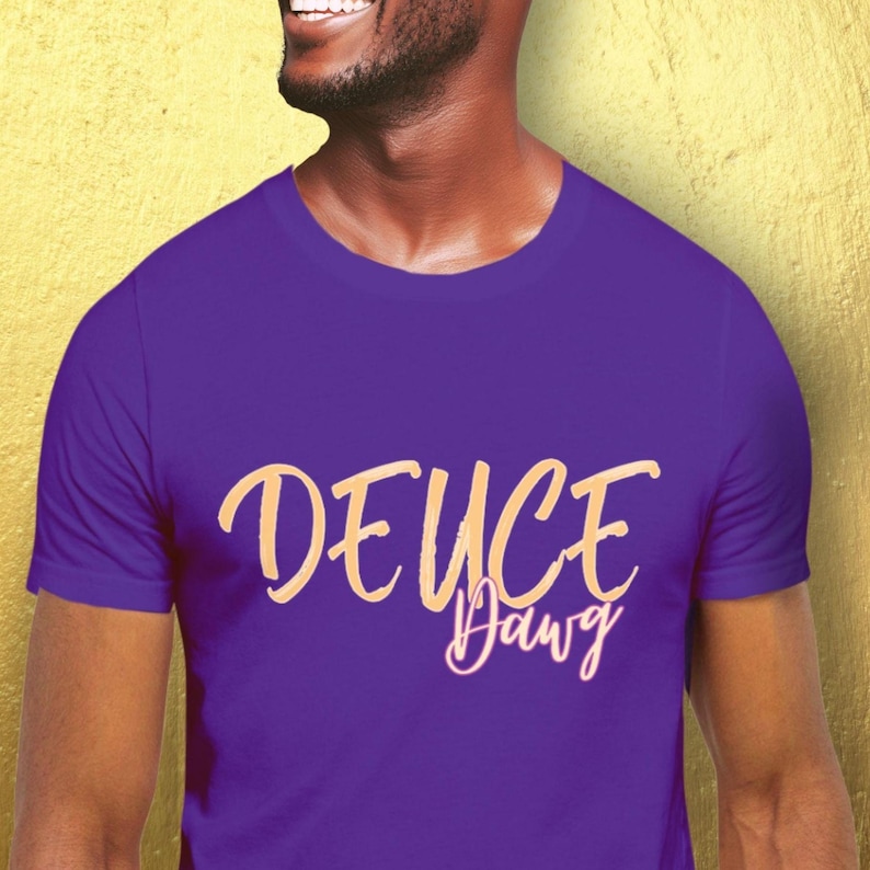 May include: A purple t-shirt featuring the text "DEUCE Dawg" in a cursive, light tan font. The shirt's solid purple colour provides a contrast to the text. The t-shirt appears to be made of a soft material.