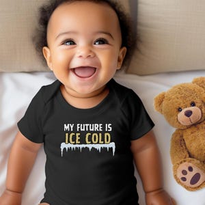 May include: A smiling baby wearing a black onesie with the text "MY FUTURE IS ICE COLD" in white and gold lettering. A brown teddy bear is next to the baby on a white surface.