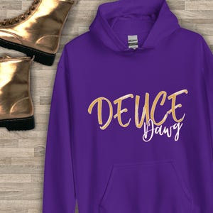 Puede incluir: Una sudadera con capucha morada con las palabras "DEUCE Dawg" en una fuente dorada. La sudadera tiene un bolsillo delantero y una capucha con cord&oacute;n. La etiqueta de la marca "Gildan" es visible en el interior de la capucha.
