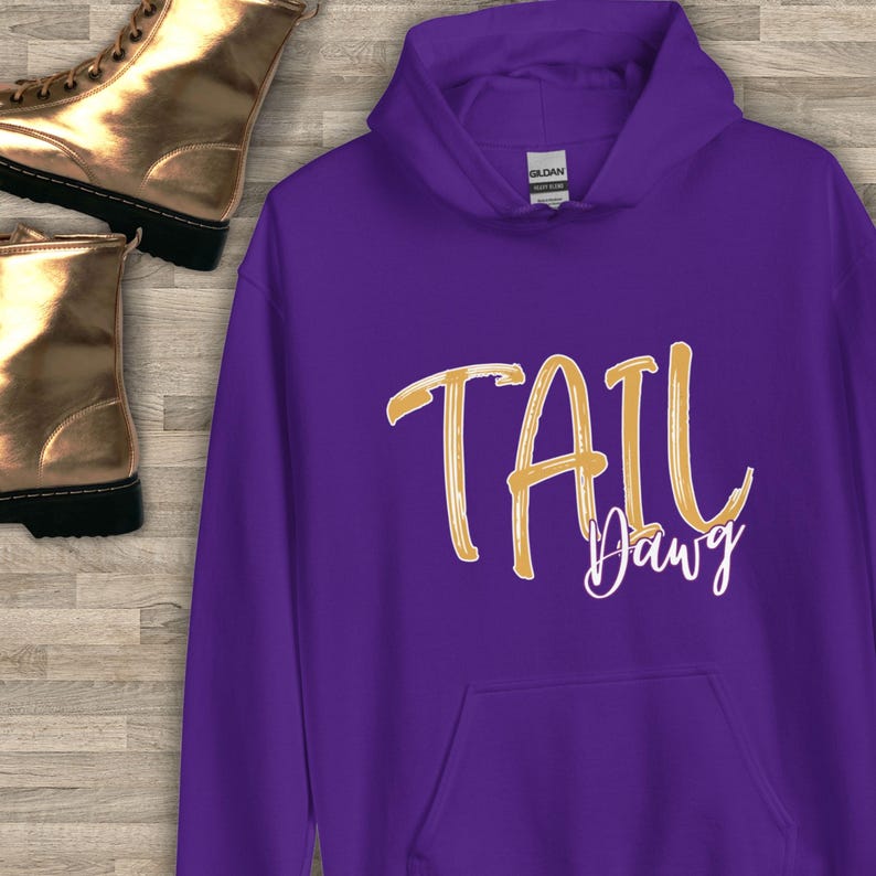 Puede incluir: Una sudadera con capucha morada con las palabras "TAIL Dawg" en letras doradas y blancas. La sudadera tiene capucha y bolsillo delantero. La etiqueta de la marca Gildan es visible.