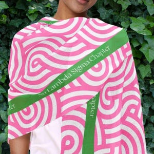 May include: A pink and white patterned scarf with a green band featuring the text "Nu Lambda Sigma Chapter". The scarf has a repeating swirl design. The scarf is draped over a white top.