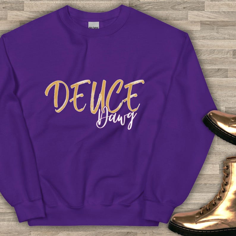 以下が含まれることがあります： 紫色のスウェットシャツ。金色の筆記体で「DEUCE Dawg」と書かれています。スウェットシャツは無地で、クルーネックです。右下には金色のブーツが見えます。