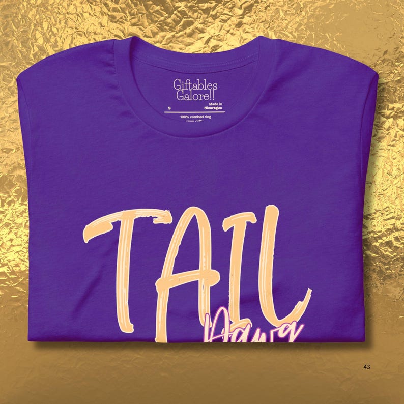 May include: A purple t-shirt featuring the word "TAIL" in a light orange script. Beneath, the word "Adura" is written in a smaller, light orange script. The t-shirt has a crew neck and short sleeves. The label reads "Giftables Galore!!" and "Made in Nicaragua".