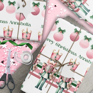 May include: Pink and green Christmas wrapping paper with a nutcracker design. The paper features a repeating pattern of pink ornaments, green bows, and nutcrackers. The text "Merry Christmas Annabella" is printed on the paper.