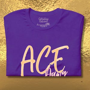 May include: A purple t-shirt featuring the word "ACE" in a light orange script. Beneath "ACE" is the word "Adair" in a smaller, pinkish font. The shirt has a crew neck and the brand "Giftables Galore!!" is printed on the tag.