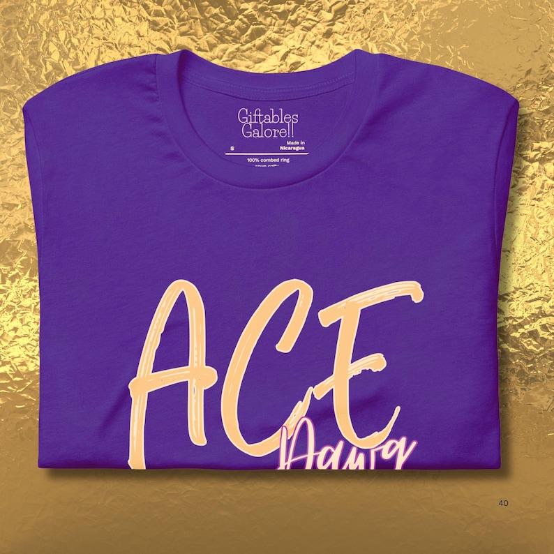 May include: A purple t-shirt featuring the word "ACE" in a light orange script. Beneath "ACE" is the word "Adair" in a smaller, pinkish font. The shirt has a crew neck and the brand "Giftables Galore!!" is printed on the tag.