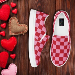 Op de afbeelding: Paar instappers met een rood-roze dambordpatroon. De schoenen hebben witte zolen en worden gepresenteerd op een houten oppervlak met hartvormige decoraties.