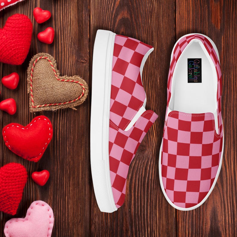 Op de afbeelding: Paar instappers met een rood-roze dambordpatroon. De schoenen hebben witte zolen en worden gepresenteerd op een houten oppervlak met hartvormige decoraties.