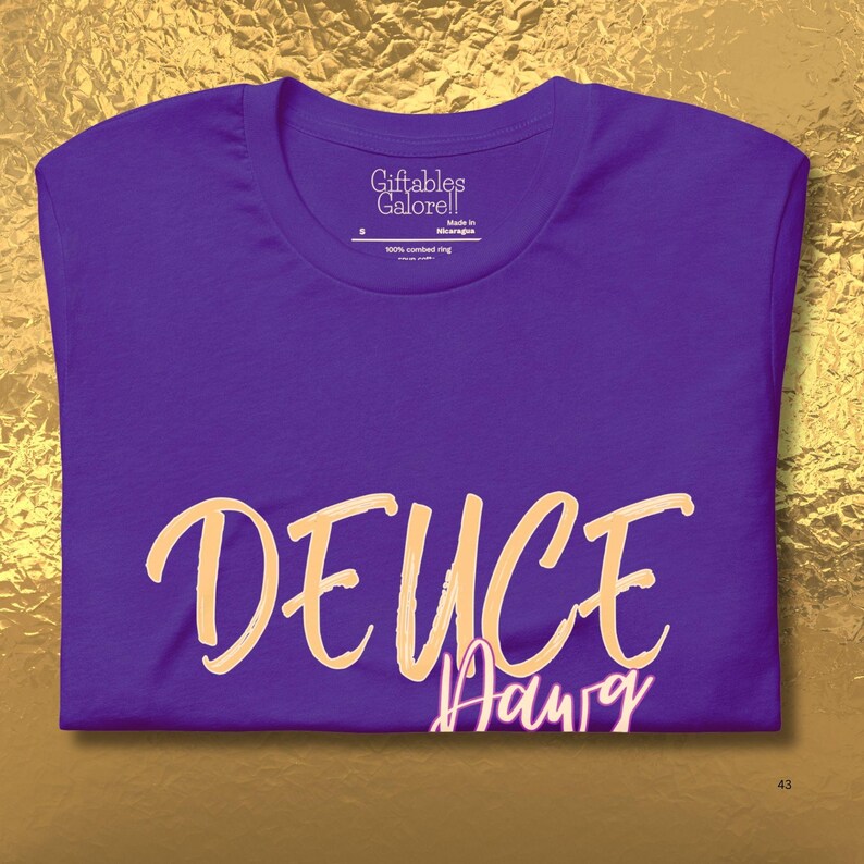 May include: A folded purple t-shirt with the word "DEUCE" in a light orange script across the front. Below the word "DEUCE" is the word "Aaurg" in a smaller, white script. The shirt has a crew neck and the tag reads "Giftables Galore!!".