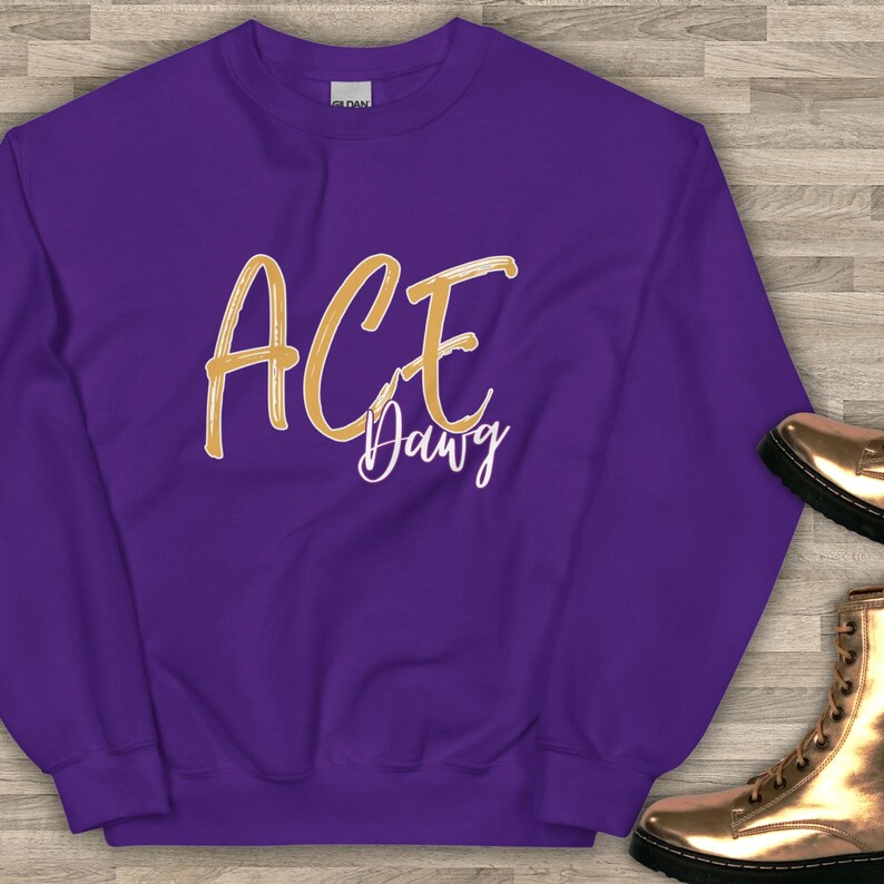 以下が含まれることがあります： 紫色のスウェットシャツ。前面には「ACE Dawg」の文字が金色の筆記体で書かれています。クルーネックと長袖のデザインです。文字はスウェットシャツの中央に配置されています。