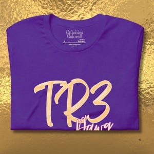 May include: A purple t-shirt featuring the text "TR3" in a light orange, brush-stroke style font. The word "Aduray" is written below in a smaller, cursive font. The shirt has a crew neck and a tag that reads "Giftables Galore!!".