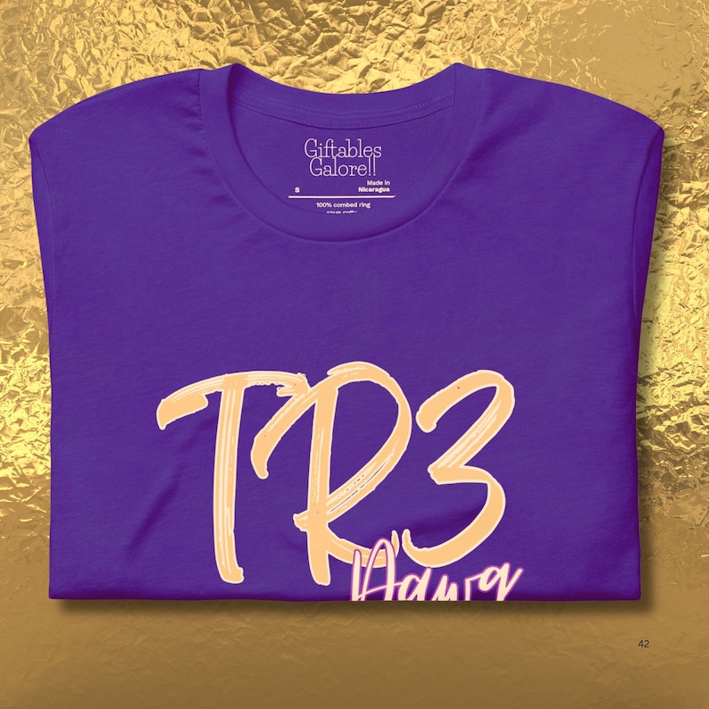 May include: A purple t-shirt featuring the text "TR3" in a light orange, brush-stroke style font. The word "Aduray" is written below in a smaller, cursive font. The shirt has a crew neck and a tag that reads "Giftables Galore!!".