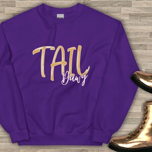 以下が含まれることがあります： 紫色のスウェットシャツ。金色の筆記体で「TAIL Dawg」と書かれています。スウェットシャツはクルーネックで、柔らかい素材でできているようです。右下には金色のブーツが見えます。