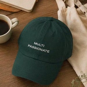May include: A dark green baseball cap with the words "MULTI PASSIONATE" embroidered in white. The cap is made of a soft material and has a curved brim. The cap is sitting on a wooden surface.