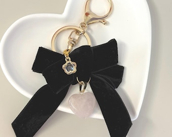 May include: A black velvet bow keychain with a gold-colored clasp and ring. A small, clear flower-shaped charm and a pink heart-shaped stone dangle from the bow. The keychain is resting on a white heart-shaped dish.