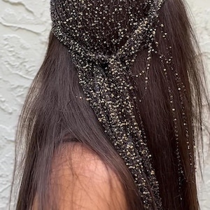 May include: A black head covering with gold sequins and beaded strands. The headpiece is worn over long, dark brown hair. The hair accessory is a decorative item, likely for special occasions or fashion.