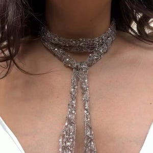May include: A silver-toned, thin, crocheted scarf or neck accessory featuring integrated small, reflective sequins throughout the open-knit mesh fabric. The accessory is wrapped around the neck and tied in a simple knot, with two long, dangling ends hanging down the chest.