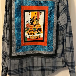 May include: A grey and black plaid button-down shirt featuring a large rectangular fabric patch on the back. The patch displays a colorful Halloween scene with the text "TRICK OR TREAT!" surrounded by orange, blue, and black fabric borders.