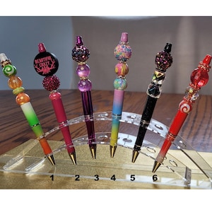 Könnte beinhalten: Sechs dekorative Stifte mit farbenfrohen, perlenbesetzten Spitzen und goldfarbenen Spitzen. Einer der Stifte trägt den Text "BEAUTIFUL CRAZY". Die Stifte werden in einem transparenten Acrylhalter präsentiert und zeigen ihre lebendigen Designs.
