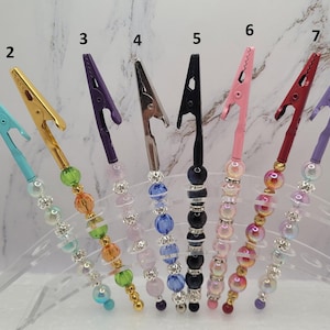 May include: Eight metal alligator clips arranged in a row, each featuring a unique handle decorated with colorful beads and rhinestones. The clips are numbered one through eight and are held in a clear acrylic stand, showcasing various metallic finishes.