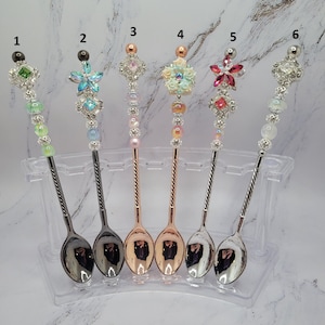 May include: Six decorative metal spoons arranged in a clear stand, numbered one through six. Each spoon features a unique handle adorned with colorful beads, rhinestones, and floral or star-shaped accents in various metallic finishes and colors.